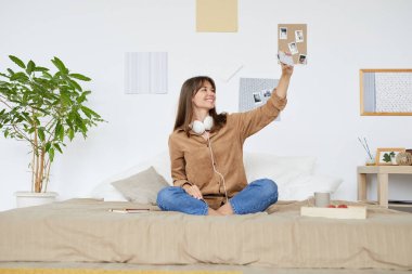 Neşeli ve çekici bir kız, günlük kıyafetleriyle yatakta bacak bacak bacak üstüne atmış ve arkadaşlarıyla internet üzerinden iletişim kurmak için mobil uygulama kullanıyor.