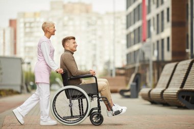 Memnuniyet verici, nitelikli bir hemşire. Engelli bir hastayla şehirde dolaşıyor. Tekerlekli sandalyeyi bir adamla itiyor.