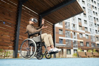 Sanal gerçeklikte tekerlekli sandalyeyi sürerken konsantrasyon gözlüklü, sokak evinin bahçesinde oturan ve el kol hareketi yapan ciddi bir genç adam.