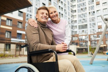 Tekerlekli sandalyeye mahkum bir adamı kucaklayan, apartmanların yakınındaki avluda yürüyen iyi kalpli bir hemşirenin portresi.