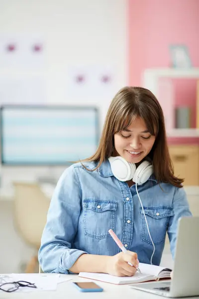 Cheerful attractive young woman in denim shirt wearing wired headphones on neck sitting at desk and writing in notepad while comparing advertising platforms