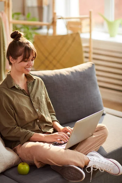 Cheerful attractive young woman with hair bun sitting with crossed legs and using laptop in own shop