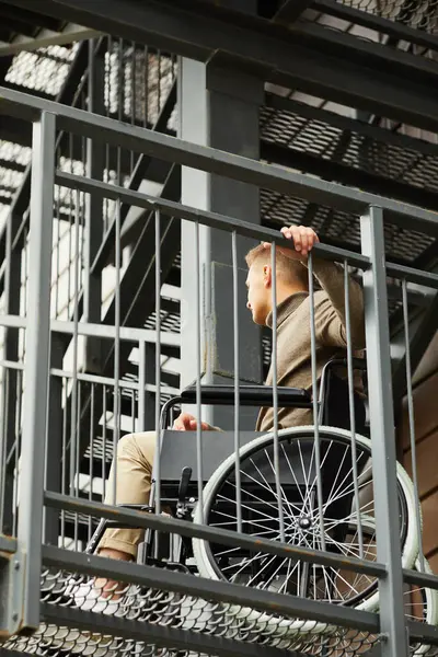 Young handicapped man in brown turtleneck sitting in wheelchair and holding metal railing while trying to go up stairs