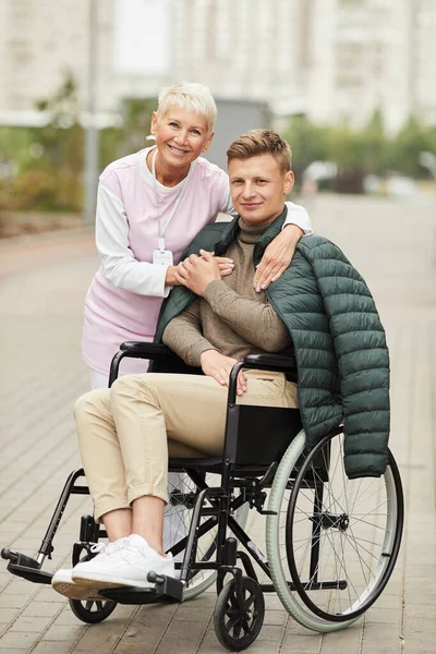 Portrait of cheerful careful female social care worker with badge spending time with patient in wheelchair outdoors