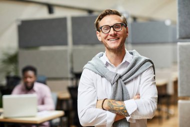 Modern ofiste kollarını kavuşturmuş, boynuna süveter dolanmış, pozitif heyecanlı genç uygulama geliştiricisinin portresi.