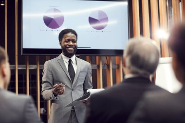 Cheerful excited young African-American sales expert in gray suit standing in front of audience and holding clipboard while presenting graphs clipart