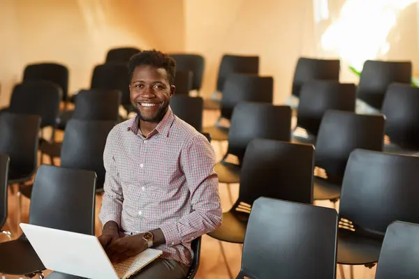 Portrait of cheerful handsome young black manager in shirt sitting on chair in conference room and using laptop