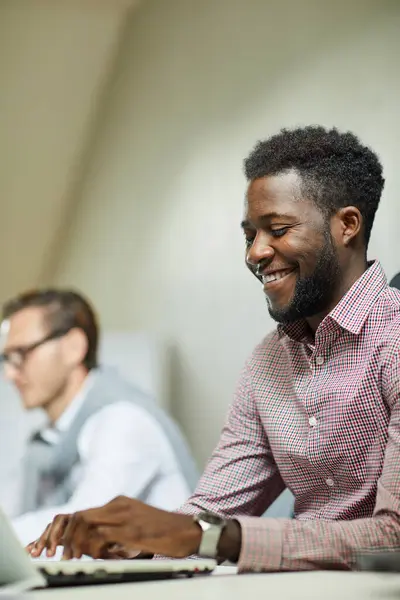 Cheerful handsome young Black programmer satisfied with web project sitting at desk in office and typing on laptop
