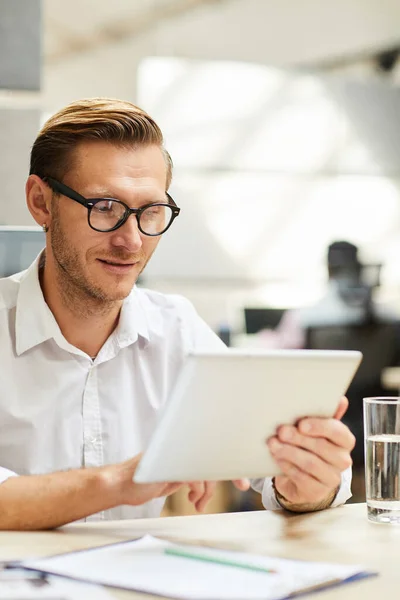 Content clever young businessman in eyeglasses sitting at table and using tablet browsing internet