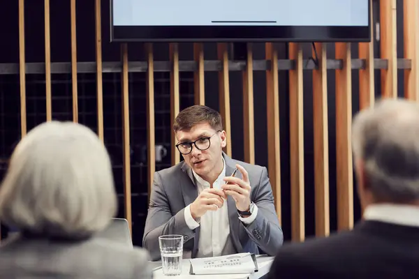 Serious smart young business expert in eyeglasses sitting at table and twisting pen while talking to colleagues at conference