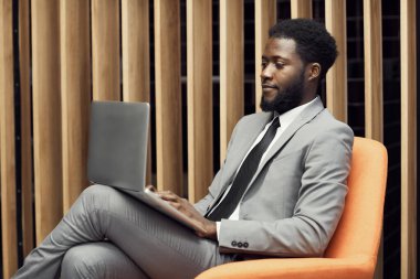 Açık gri takım elbiseli ciddi sakallı Afro-Amerikalı yönetici rahat koltukta oturuyor ve dizüstü bilgisayarında yazıyor.