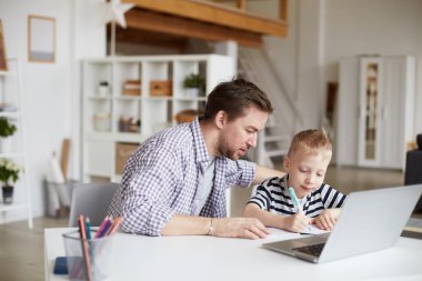 Ciddi bir genç baba. Günlük gömlekli. Masada dizüstü bilgisayarla oturuyor ve oğlunun ödevlerini yapmasına yardım ediyor.