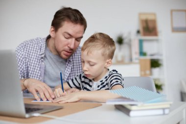 Ciddi, genç bir baba, odada oturmuş, oğlunun çalışma kitabını yazdığı iş çözümünü inceliyor.