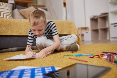 Sarı saçlı, günlük kıyafetli, çapraz bacaklı, kalemli ve oturma odasındaki kağıda çizim yapan meşgul bir çocuk.