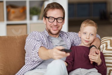 Üzgün sarı saçlı oğlan babasının TV kanalını seçmesinden memnun değil. Kanepede oturup babasıyla televizyon izliyor.