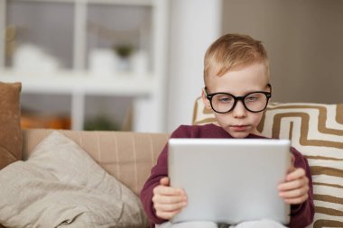 Gerçekten meraklı küçük bir çocuk kanepeye oturmuş online kitap okurken dijital tablet kullanıyor.