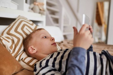 İnternette film izlerken yatağa uzanan ve dijital tablet kullanan ciddi bağımlı çocuğun yakın çekimi.