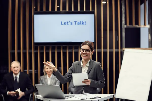 Portrait of positive excited young business lady in eyeglasses standing at speech table and producing arguments while giving speech at meeting