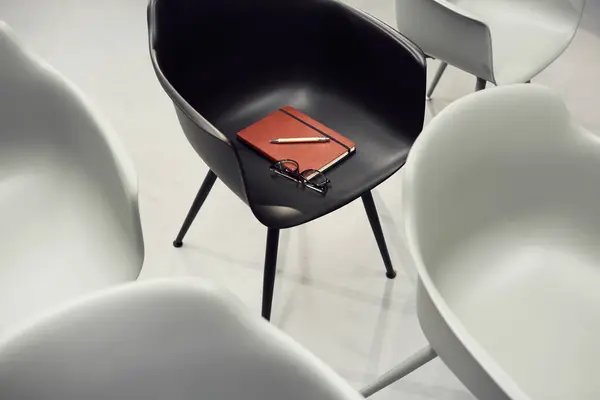 Red notepad, eyeglasses and pen on black chair among white seats in conference room