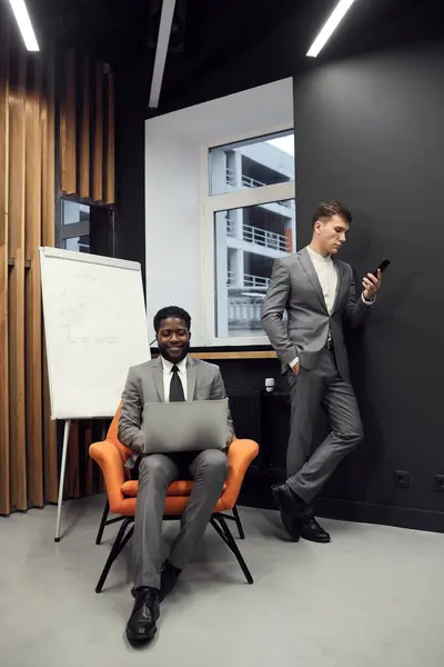 Young successful managers working with social medias: smiling black businessman sitting in orange armchair and typing on laptop while his colleague scrolling page on internet using smartphone