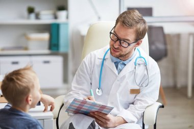 Beyaz önlüklü nazik genç doktor küçük bir hastayla semptomlar hakkında konuşuyor ve tıbbi kartıyla notlar alıyor.