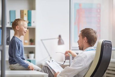 Gülümseyen sarı saçlı çocuk muayene masasında oturmuş neşeyle doktorla konuşuyor.