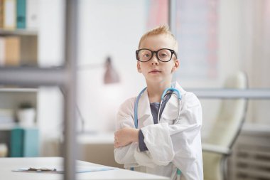 Laboratuvar önlüklü ve gözlüklü, klinik odasında ayakta duran ve kollarını göğsünde kavuşturan kendine güvenen küçük bir doktorun portresi.