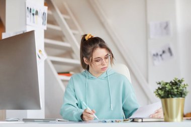Gözlüklü ve kapüşonlu ciddi bir genç kadın masada oturmuş web projesi için skeçleri analiz ediyor.
