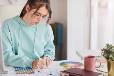Kapüşonlu ciddi bir kadın tasarımcı masada oturmuş kalemle moda skeci çiziyor.