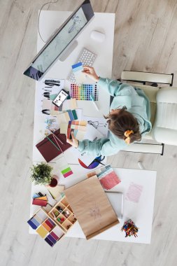 Kapüşonlu genç bir kadının yeni elbiseler için renk seçerken masasında oturup paletlere baktığı yüksek açı.