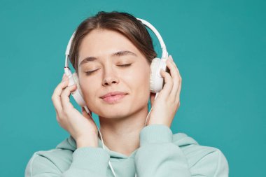 Sakin, çekici, gözleri kapalı, kulaklık takan, deniz mavisi arka planda dinlenen müzik dinleyen genç bir kadın.