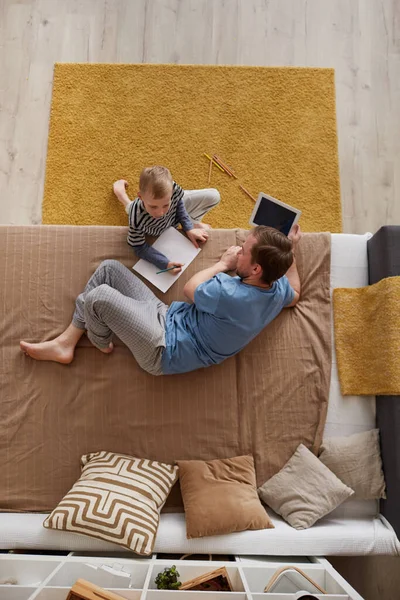Above view of little blond-haired boy sitting at sofa bed and making picture with father who surfing internet at tablet
