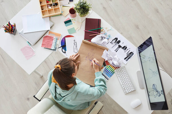 Высокий угол обзора молодой женщины в толстовке, сидящей за столом с палитрами и компьютером и рисующей эскизы модной одежды в студии