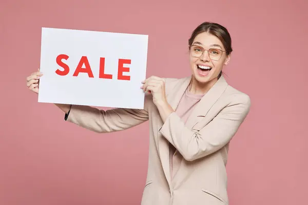 戴着眼镜的快乐而美丽的年轻女性对站在粉色背景下展示销售海报感到兴奋 — 图库照片