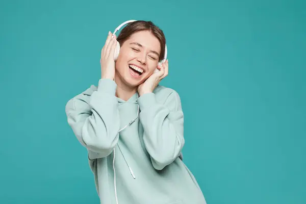 Cheerful ecstatic young woman in hoodie keeping eyes closed and laughing while enjoying music in headphones, studio shot