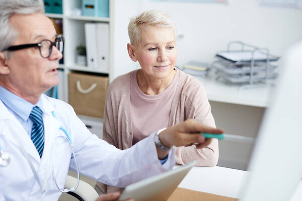 Serious senior male doctor sitting at table and pointing at monitor while explaining CAT scan to senior patient
