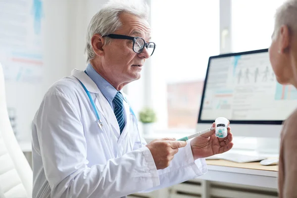 Serious wrinkled physician in glasses pointing with pen at pill bottle while advising patient to take medicine