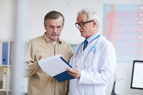 Serious senior doctor in lab coat and glasses holding clipboard with medical papers and watching test results with aged patient