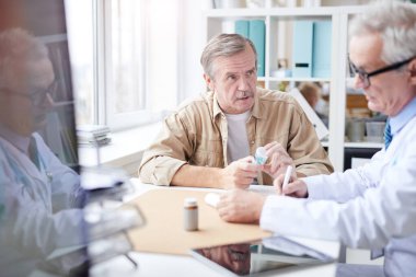 Ciddi derecede endişeli bıyıklı son sınıf öğrencisi masada oturmuş, elinde ilaç şişesiyle doktora yeni ilaçlarını soruyor.