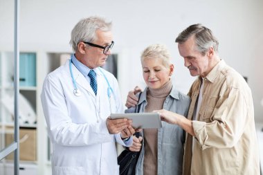 Steteskopu boynunda olan yakışıklı bir doktor dijital tablet kullanırken kocasıyla birlikte iyileşen hastasına yararlı bilgiler veriyor.