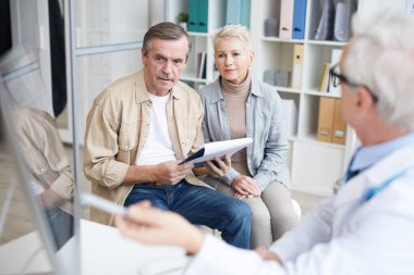 Doktor muayenehanesinde oturan ve doktorları dinlerken reçete okuyan karısı tarafından desteklenen bıyıklı kıdemli hasta.