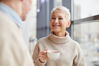 Gülümseyen, kısa saçlı, çekici, olgun bir bayan. Pencerede duruyor ve arkadaşınla konuşurken çay içiyor.