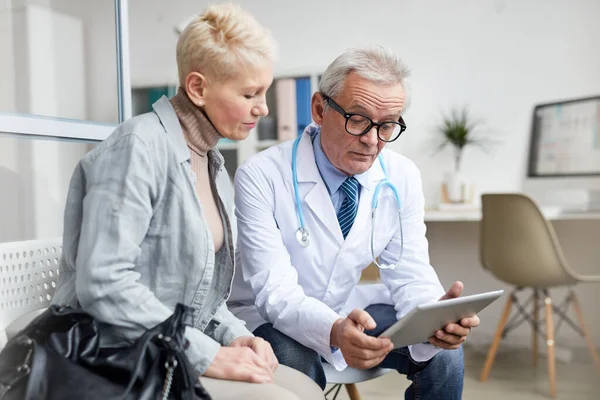 Serious attractive mature woman in casual outfit sitting on chair in doctors office and listening to doctor using tablet to analyze test results during appointment