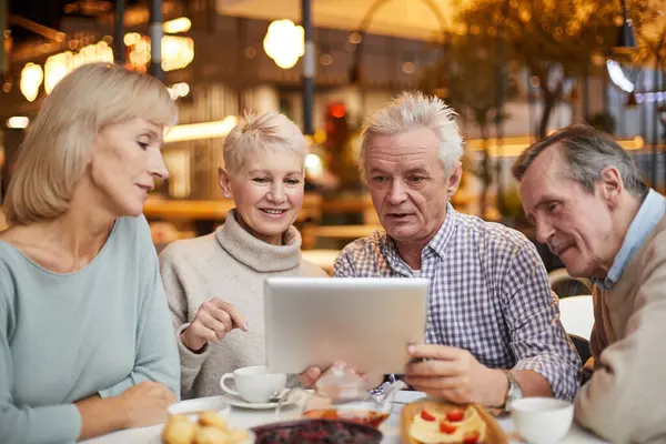Group of content curious senior friends in casual outfits sitting at table in cafe with wi-fi and surfing net on tablet together