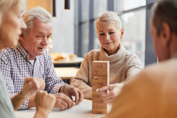 Group of content concentrated senior people sitting at table in nursing home and playing jenga together