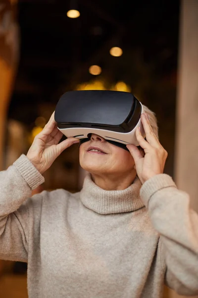 Smiling mature woman in turtleneck sweater holding virtual reality glasses while watching 3D video