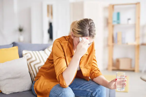 Depressed girl in orange blouse sitting on sofa and drinking water while discussing her heartbreak with psychologist