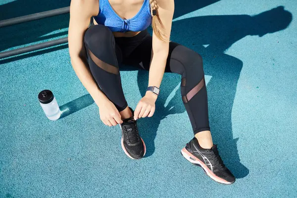 Close-up of unrecognizable woman in leggings sitting on ground and tying shoe while preparing for outdoor training