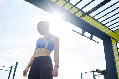 Güneş ışığında egzersiz alanında duran, mükemmel vücut şekilleri olan, ciddi, güzel, sarışın bir kadın.