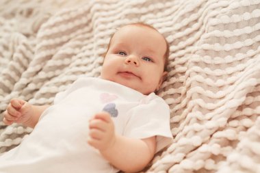 Beyaz pamuklu tulumlu küçük bebek yatakta uzanıyor ve kameraya gülümsüyor.